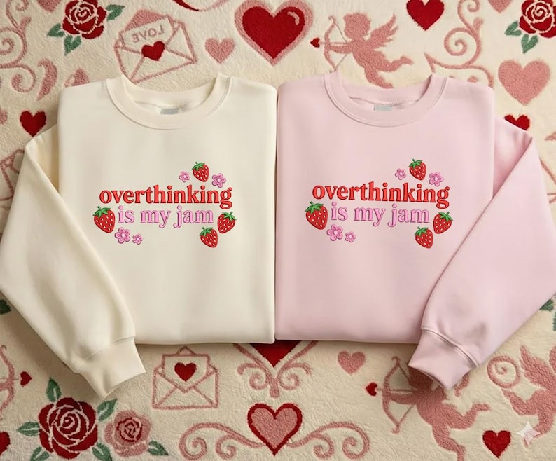 May include: Two crewneck sweatshirts, one cream and one pink, with the phrase "overthinking is my jam" embroidered in red. The design includes strawberries and flowers. The sweatshirts are displayed on a patterned surface with hearts and roses.