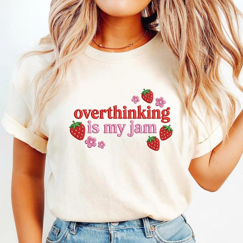 May include: Cream-colored t-shirt with the phrase "overthinking is my jam" in red and pink lettering. The design includes strawberry and flower graphics. The shirt has rolled-up sleeves.
