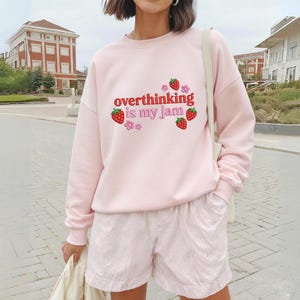 May include: A light pink sweatshirt with the phrase "overthinking is my jam" in red script, accented with strawberry and flower graphics. The model is also wearing pink shorts.