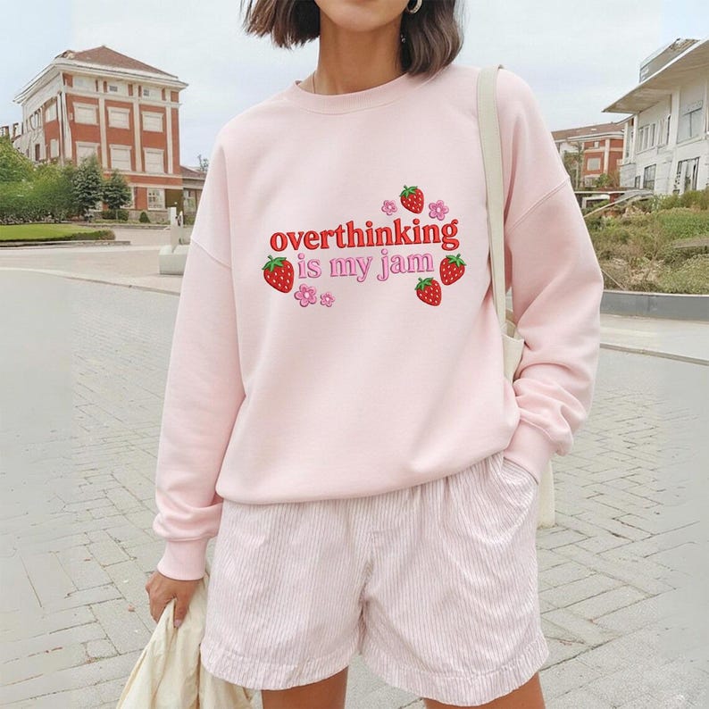 May include: A light pink sweatshirt with the phrase "overthinking is my jam" in red script, accented with strawberry and flower graphics. The model is also wearing pink shorts.