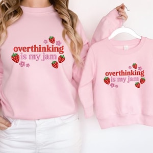 May include: Two matching light pink sweatshirts. The adult sweatshirt is worn by a person, and the child's sweatshirt is held up. Both sweatshirts have the text "overthinking is my jam" in red, with strawberry and flower graphics.