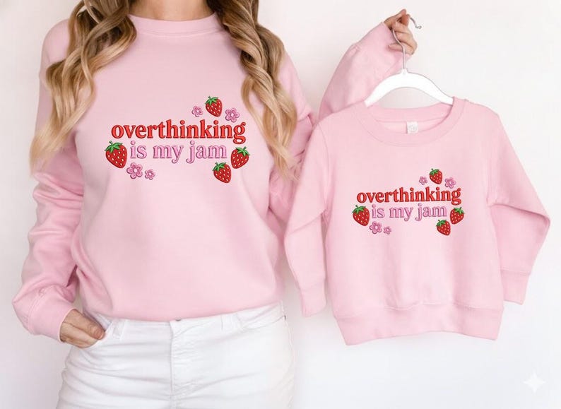 May include: Two matching light pink sweatshirts. The adult sweatshirt is worn by a person, and the child's sweatshirt is held up. Both sweatshirts have the text "overthinking is my jam" in red, with strawberry and flower graphics.