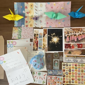 Könnte beinhalten: Eine Sammlung japanischer Schreibwaren, darunter gemustertes Origamipapier, vier gefaltete Papierkraniche in Gelb, Lila, Grün und Blau, Postkarten mit Festivalszenen, Aufkleberbögen mit Essensmotiven, ein kleiner Faltfächer und eine handgeschriebene Notiz.