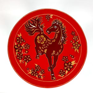 May include: A round, red decorative piece featuring a prancing horse design. The horse is a deep burgundy color with gold accents, surrounded by gold floral patterns. The background is a vibrant red.