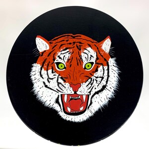 May include: Round black decorative art piece featuring a tiger's face. The tiger has orange and white fur with green eyes and a red mouth. The tiger is roaring, with its teeth visible. The art piece is likely a painting or print.