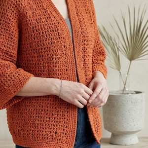 Pode incluir: Cardigã de crochê cor ferrugem com mangas três-quartos. O cardigã aberto tem um design de tecido aberto. A modelo usa-o sobre um top cinzento e calças de ganga azuis. Uma pulseira prateada é visível no pulso da modelo.