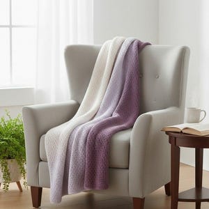 Op de afbeelding: Een zachte, gebreide deken over een grijze fauteuil. De deken heeft een kleurovergang van wit naar lavendel naar diep paars. De stoel staat in een lichte kamer met een bijzettafel en een plant in een pot.