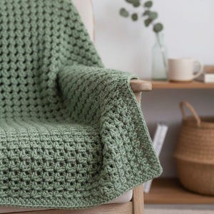 May include: A sage green crocheted blanket draped over a wooden chair. The blanket has a textured, open weave pattern. The chair has a light-colored cushion and wooden armrest. The blanket's edge is visible, showing the detailed stitch work.
