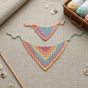 May include: Two pastel-colored, crocheted bandanas. The larger one is in the foreground, with a rainbow of colors including pink, yellow, and blue. The smaller one is above it. Knitting supplies, including yarn and buttons, are in the background.
