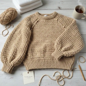 May include: A tan, hand-crocheted sweater with a textured stitch pattern and balloon sleeves. The sweater has a ribbed hem and cuffs, and wooden buttons at the shoulder. A ball of yarn, crochet hook, and a tag that says "Cloudline Sweater - Handmade" are also visible.