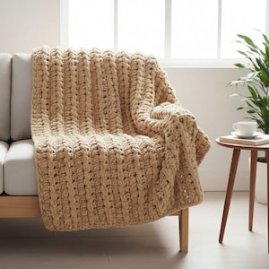 Puede incluir: Una manta beige de punto grueso sobre un sofá de color claro. La manta tiene un tejido grueso y texturizado, creando una apariencia acogedora y atractiva. El color neutro complementa la decoración de la habitación. La manta mide aproximadamente 152 cm de largo.