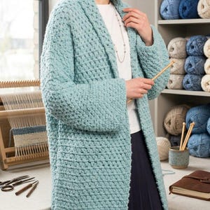 Könnte beinhalten: Hellblauer, grob gestrickter Cardigan mit Schalkragen. Der lange, offen geschnittene Pullover ist aus dickem, strukturiertem Garn gefertigt. Stricknadeln und Wollknäuel sind im Hintergrund zu sehen, was auf ein handgefertigtes Handwerk hindeutet.