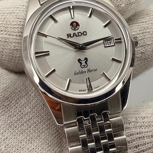 May include: Silver Rado Golden Horse wristwatch with a silver face, black hands, and a date window at the 3 o'clock position. The watch has a stainless steel strap and the Rado logo. A classic timepiece.