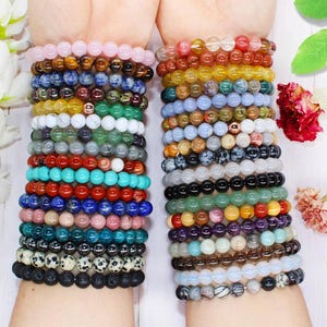 Könnte beinhalten: Eine Sammlung von Perlenarmbändern in verschiedenen Farben, darunter Rosa, Braun, Blau, Grün, Rot und Schwarz. Die Armbänder bestehen aus runden Perlen und sind an Handgelenken gestapelt. Die Armbänder werden auf einer weißen Oberfläche präsentiert.
