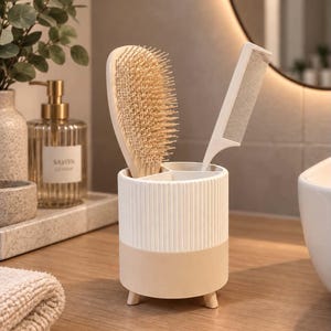 May include: A cylindrical bathroom organiser with a ribbed white upper section and a smooth beige base, supported by three small feet. It holds a beige hairbrush with flexible bristles and a white plastic hair comb in its divided interior compartments.
