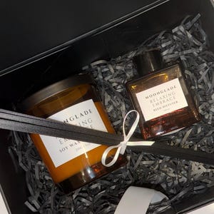 May include: A gift box containing a candle and reed diffuser. The candle is in a glass jar with a dark lid, and the diffuser is in a square bottle. Both have labels that read "MOONGLADE Relaxing Embrace". The box is filled with shredded paper.