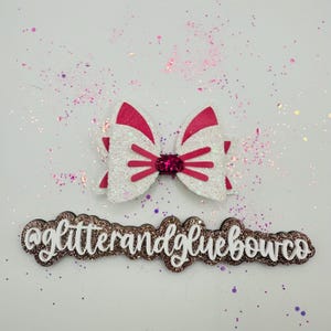 以下が含まれることがあります： ピンクのアクセントとピンクの中央部を備えた白いグリッターリボン。リボンはピンクの線で装飾され、散らばったグリッターのある白い背景に配置されています。テキスト「@glitterandgluebowco」はリボンの下にあります。