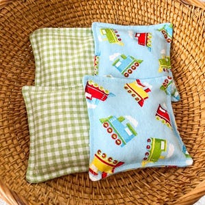 May include: A woven basket holds three small, square fabric items. Two are light blue with colorful train illustrations, and one is green and white gingham. The basket is made of woven brown material.