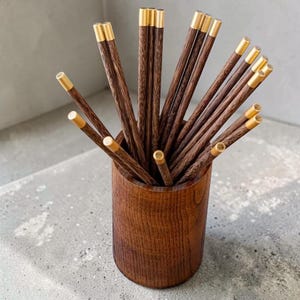 May include: A collection of wooden chopsticks with gold-colored tips, displayed in a cylindrical wooden holder. The chopsticks have a dark brown color with visible wood grain. The holder is also made of wood, with a similar dark brown finish.