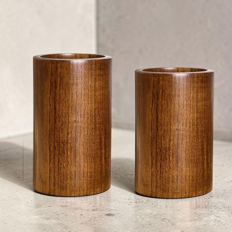 May include: Two cylindrical wooden containers with a rich brown finish. The containers are of different sizes, both with a hollowed-out interior. The wood grain is visible, adding texture to the smooth, rounded forms. They sit on a light-colored surface.