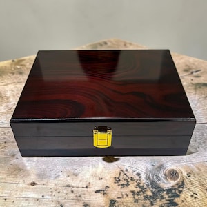 May include: A rectangular wooden box with a dark, polished mahogany finish featuring a prominent wood grain pattern. The box is closed and secured with a small, centered gold-colored metal latch on the front edge, providing a secure closure.