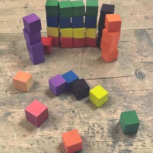Könnte beinhalten: Eine Sammlung kleiner, einfarbiger Holzwürfel in verschiedenen Farbtönen wie Rot, Orange, Gelb, Grün, Blau, Lila und Schwarz. Einige Würfel sind zu vertikalen Türmen gestapelt, während andere einzeln auf der Oberfläche verstreut liegen.