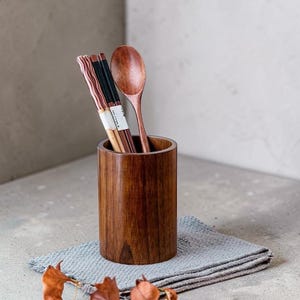May include: A wooden utensil holder with a set of chopsticks and a wooden spoon. The holder is cylindrical and made of dark brown wood. The chopsticks are black and brown, and the spoon is a lighter shade of brown. The holder sits on a gray cloth.