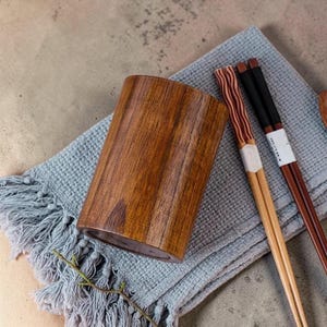 May include: A wooden utensil holder with a natural wood grain finish. Two pairs of chopsticks rest on a blue fringed cloth. One pair has a black handle, and the other has a red and brown striped design.
