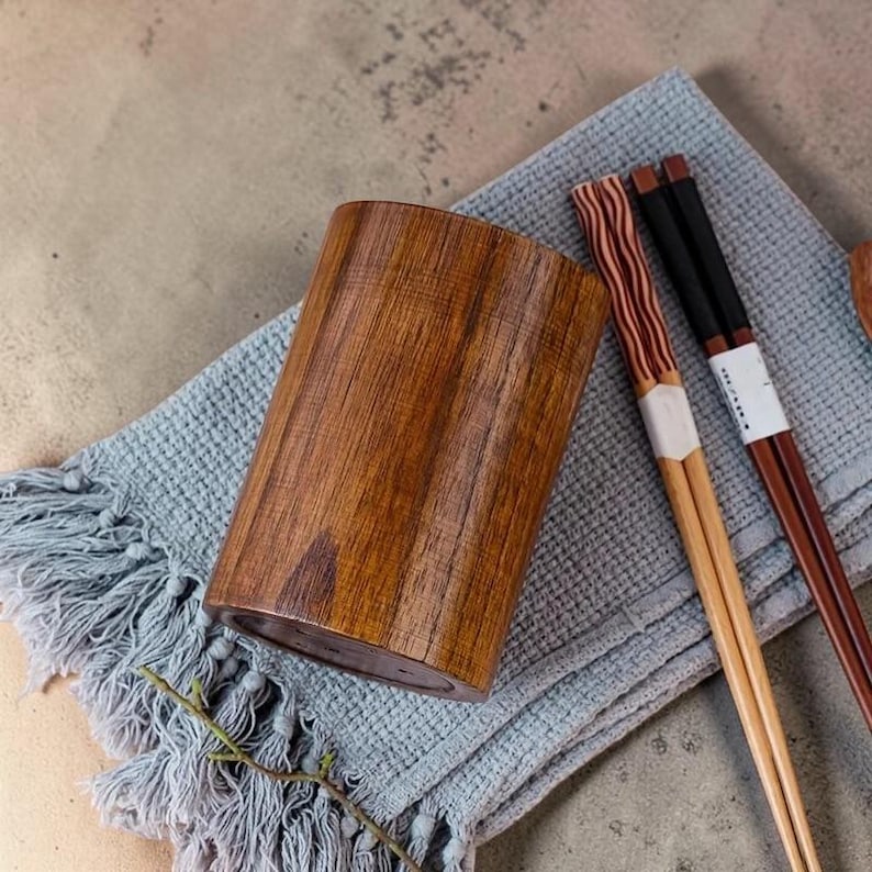 May include: A wooden utensil holder with a natural wood grain finish. Two pairs of chopsticks rest on a blue fringed cloth. One pair has a black handle, and the other has a red and brown striped design.
