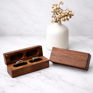 Könnte beinhalten: Eine dunkelbraune Holzringbox mit zwei Fächern, eines davon mit einem silbernen Ring. Die Box ist geöffnet und zeigt ein schwarzes Samtfutter. Sie steht auf einer Marmoroberfläche.