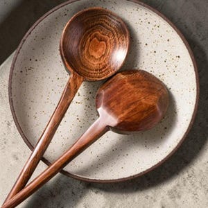 Könnte beinhalten: Eine weiße Keramik-Tasse mit schwarzem Henkel und dem Text "But First, Coffee" in schwarzer, handschriftlicher Schrift. Die Tasse steht auf einer weißen Oberfläche.