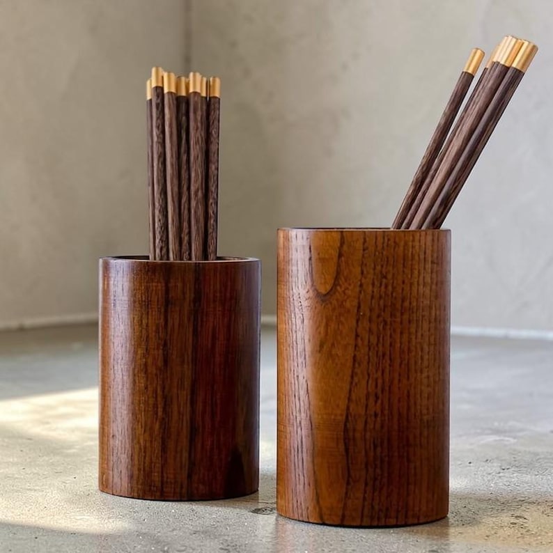 May include: Two wooden chopstick holders with chopsticks. The holders are cylindrical and made of dark brown wood. The chopsticks are dark brown with gold-colored tips. The holders are on a light gray surface.