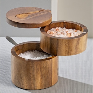 Könnte beinhalten: Ein zweistöckiger Salz- und Gewürzbehälter aus Holz mit Deckel und kleinem Löffel. Das obere Fach enthält rosa Himalaya-Salz, während das untere weißes Salz enthält. Das Holzdesign verleiht der Küche einen rustikalen Touch.