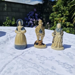 May include: Three ceramic figurines standing on a white tablecloth. The left figure wears a blue bonnet and dress. The centre figure is a person in a tan coat and hat with a dog. The right figure wears a blue headscarf and tan dress with the name KATHLEEN.