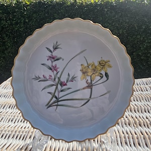 May include: A round white ceramic quiche dish with a scalloped edge and a thin gold rim. The centre of the dish features a botanical illustration of yellow daffodils and small pink flowers with green stems and leaves on a plain white background.