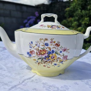 Op de afbeelding: Een witte keramische theepot met een lichtgele rand aan de bovenkant en de basis. Het lichaam is versierd met een kleurrijk boeket van rode, blauwe, gele en roze bloemen. De theepot is voorzien van een bijpassend deksel en een gebogen tuit.