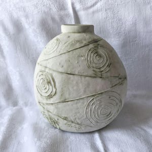 Może przedstawiać: Okrągły, kremowobiały wazon ceramiczny o teksturowanej, matowej powierzchni. Zewnętrzna strona posiada wyryte wzory spiralne i ukośne linie, z subtelnymi zielonkawymi przebarwieniami lub śladami w zagłębieniach i na powierzchni.