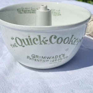 Puede incluir: Un molde para pudin de cerámica blanca con una chimenea central, con texto en verde que dice The Quick-Cooker y Grimwade's Patent No 12835/09. El borde interior incluye instrucciones escritas sobre el llenado para la expansión del pudin.