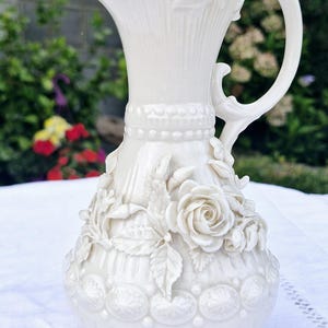 May include: A cream-coloured ceramic pitcher featuring an ornate relief design of blooming roses and leaves. The vessel has a flared rim, a curved handle, and a decorative band of raised circular patterns around the neck and base of the body.