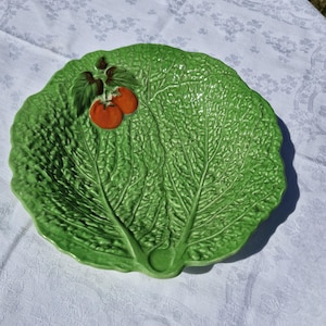 Puede incluir: Un plato redondo de cerámica verde con un patrón texturizado y en relieve de hoja de col en toda su superficie. Dos pequeños tomates de color rojo anaranjado con hojas de color verde oscuro están fijados en la parte superior izquierda.