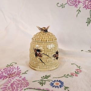 Puede incluir: Un recipiente de cerámica con forma de colmena, con tapa, que presenta una textura tejida detallada y adornado con dos abejas pintadas en vuelo. Una sola abeja descansa sobre la tapa. El recipiente es de color amarillo verdoso claro.