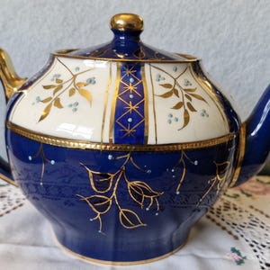 Op de afbeelding: Een kobaltblauwe en crèmekleurige theepot met gouden bladmotieven, een goudkleurig handvat en een bijpassend deksel met een gouden knop. De behuizing is voorzien van gedetailleerd goudfiligraan, kleine blauwe stippen en een centraal verticaal blauw paneel.