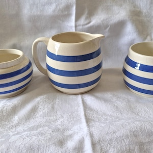 Peut inclure: Un ensemble en céramique composé d'un pichet et de deux bols assortis, tous dotés d'une base couleur crème avec des rayures horizontales bleu marine. Le pichet possède une anse incurvée et un bec verseur, tandis que les bols sont arrondis.
