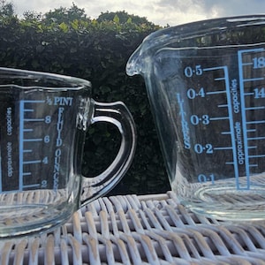 Op de afbeelding: Twee doorzichtige glazen maatbekers met blauwe markeringen. De kleinere beker toont maten in liters en vloeibare ounces tot 0,3 liter en 8 vloeibare ounces. De grotere beker toont maten tot 0,5 liter en 18 vloeibare ounces.