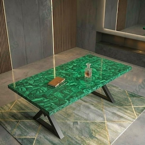 Può includere: Un tavolino rettangolare caratterizzato da una superficie con motivo in malachite verde brillante e finitura lucida. Il tavolo è sostenuto da una struttura in metallo nero con gambe angolate. Un piccolo oggetto rettangolare marrone e un decanter in vetro sono appoggiati sopra.