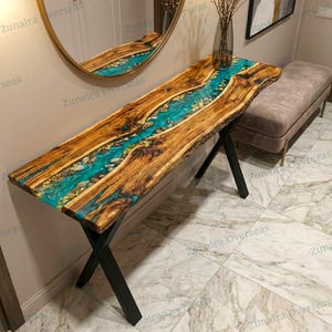 Puede incluir: Una consola de madera con un diseño de río de resina turquesa. La mesa presenta un borde natural, vetas de madera natural y patas de metal negro. Acentos de pan de oro están incrustados en la resina. La mesa mide aproximadamente 152 cm de largo.