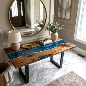 May include: A wooden console table with a blue resin river design. The table has a natural wood grain finish and black metal legs. A gold-framed mirror hangs above the table, reflecting the room. The table is 60 inches long.
