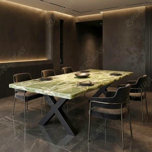 Puede incluir: Una mesa de comedor rectangular con una superficie de piedra ónix verde pulida con vetas blancas y una base de metal negro en forma de X. La mesa está rodeada por seis sillones tapizados en gris oscuro con marcos de metal delgados.