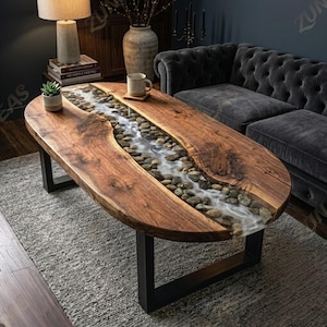 May include: Oval wooden coffee table with a river-like design. The table features a dark wood surface with a clear resin river filled with stones. It has a black metal base and is placed on a gray rug.