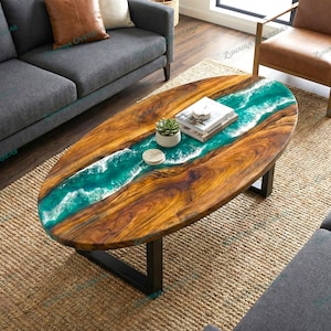 Op de afbeelding: Ovale salontafel van hout met een turquoise hars rivierontwerp. De tafel heeft een donkere houtnerf afwerking en zwarte metalen poten. Een kleine plant en boeken liggen op de tafel. De tafel staat op een geweven tapijt.